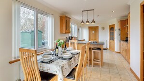Une cuisine spacieuse avec des meubles en bois, un coin repas avec une table dressée pour quatre personnes, et une vue sur l’extérieur grâce à de grandes fenêtres.