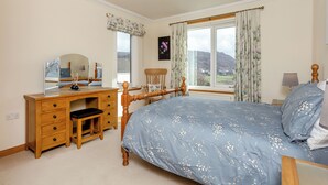 Une chambre à coucher comprenant un lit, une commode en bois, un bureau, une chaise, un miroir, une fenêtre avec des rideaux et une vue sur l’extérieur.
