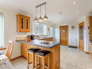 A kitchen with wooden cabinets, a central island with stools, pendant lighting, a window, and a radiator. - Welcome to Bramble Cottage. (Gairloch)