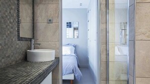 A modern bathroom with a round sink, a glass shower enclosure, and a visible bed in the adjacent room.