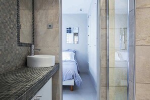 A modern bathroom with a round sink, a glass shower enclosure, and a visible bed in the adjacent room.