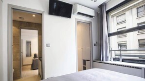 A hotel room with a bed, a TV mounted on the wall, an air conditioning unit, and a window with a view of the building outside.