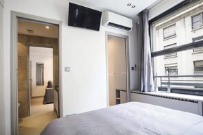 A hotel room with a bed, a TV mounted on the wall, an air conditioning unit, and a window with a view of the building outside.
