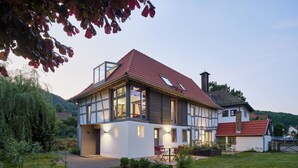 Une maison moderne à deux étages, avec un toit recouvert de tuiles rouges, de grandes fenêtres et un jardin bien entretenu agrémenté de pierres plates servant de pas japonais.