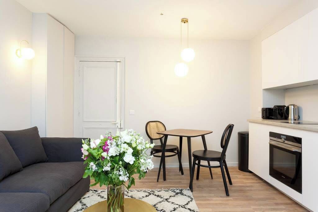 A modern living room with a sofa, a dining table with chairs, a vase of flowers, and a kitchen area with a stove and coffee maker.