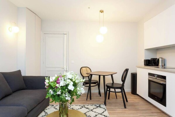 Un salon moderne comprenant un canapé, une table à manger avec des chaises, un vase de fleurs et un coin cuisine avec une cuisinière et une machine à café.