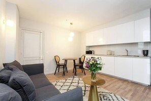 A modern living room with a sofa, a dining table with chairs, a kitchen area with white cabinets, and a vase with flowers on a table.