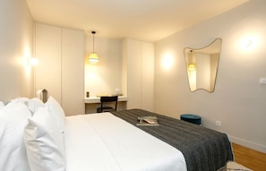 A hotel room with a bed, a desk, a chair, a mirror, and a small ottoman.