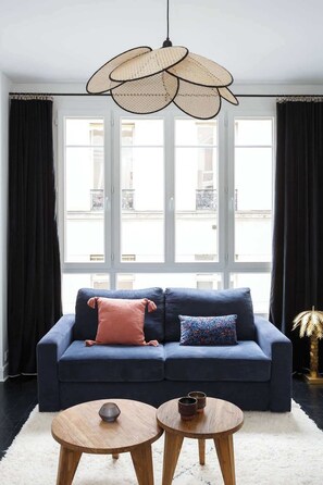 Uma sala de estar moderna com um sofá azul escuro, duas mesas redondas de madeira, uma luminária pendente trançada e grandes janelas com cortinas pretas.
