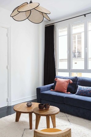 A living room with a dark blue sofa, two round wooden tables, a woven pendant light, and a view through large windows.