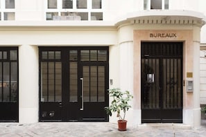 A building with a 'BUREAUX' sign, featuring large black doors with glass panels, a potted plant in front, and a stone facade.