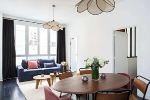 A modern living room with a dark blue sofa, a round wooden dining table, and a large window with black curtains.