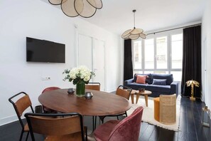 A modern living room with a round wooden dining table, a flat-screen TV mounted on the wall, and a dark-colored sofa with cushions.