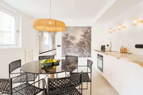 A modern kitchen with a round dining table, black wicker chairs, a hanging light fixture, and a tropical mural on the wall.