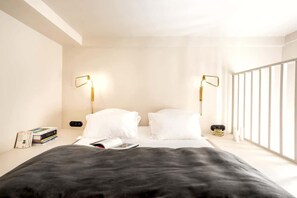 A modern bedroom with a large bed, two bedside lamps, a bedside table with books, and a window with blinds.