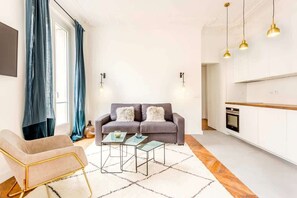 Una sala moderna con un sofá gris, una mesa de centro de vidrio y un sillón con marco dorado. Hay una zona de cocina con armarios blancos y un horno empotrado.