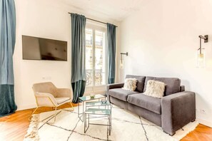 A living room with a grey sofa, a glass coffee table, a beige armchair, and a TV mounted on the wall. - Elegant spacious apartment with balcony near Opéra - 2 bedrooms - A/C - Steps from the grands magasins - Quatre Septembre by Habitat Parisien (Paris)