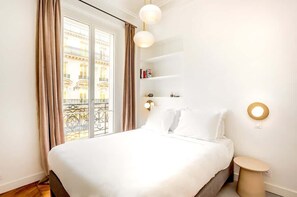 Habitación de hotel con una cama grande, una mesita de noche pequeña, un estante en la pared con libros y una ventana con cortinas.