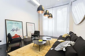 Uma sala de estar moderna com um sofá preto, uma mesa de centro redonda e duas cadeiras pretas. Há duas luminárias pendentes no teto e um pôster grande na parede.