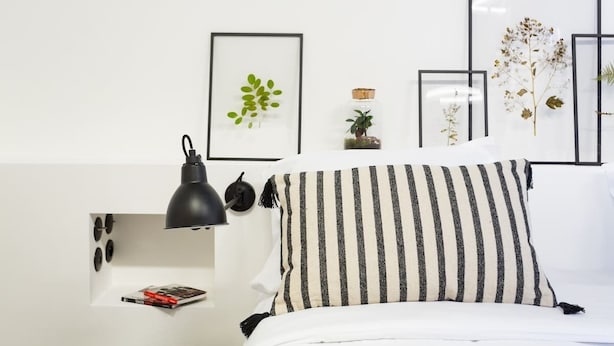 A neatly arranged bedroom with a bed, a bedside lamp, a framed plant, and a small shelf with a book.