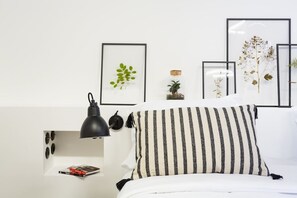 Un dormitorio ordenado con cama, lámpara de noche, planta enmarco y una pequeña estantería con un libro.