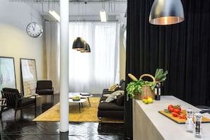 A modern living room with a sofa, chairs, a coffee table, and a kitchen area with a cutting board, vegetables, and a bottle.