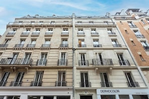 A multi-story building with a classic architectural style, featuring balconies with wrought iron railings and a storefront with a sign reading "DERMOCOLOR".