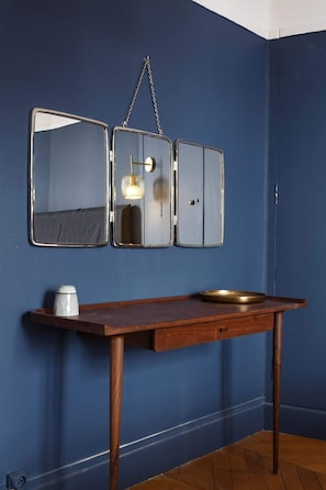 A room with a blue wall, a wooden table with a metal bowl, and a wall-mounted mirror with a hanging light fixture.