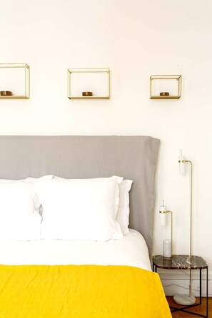 Un lit bien fait, avec du linge de lit blanc et un plaid jaune, une table de chevet avec une lampe, et trois appliques murales.