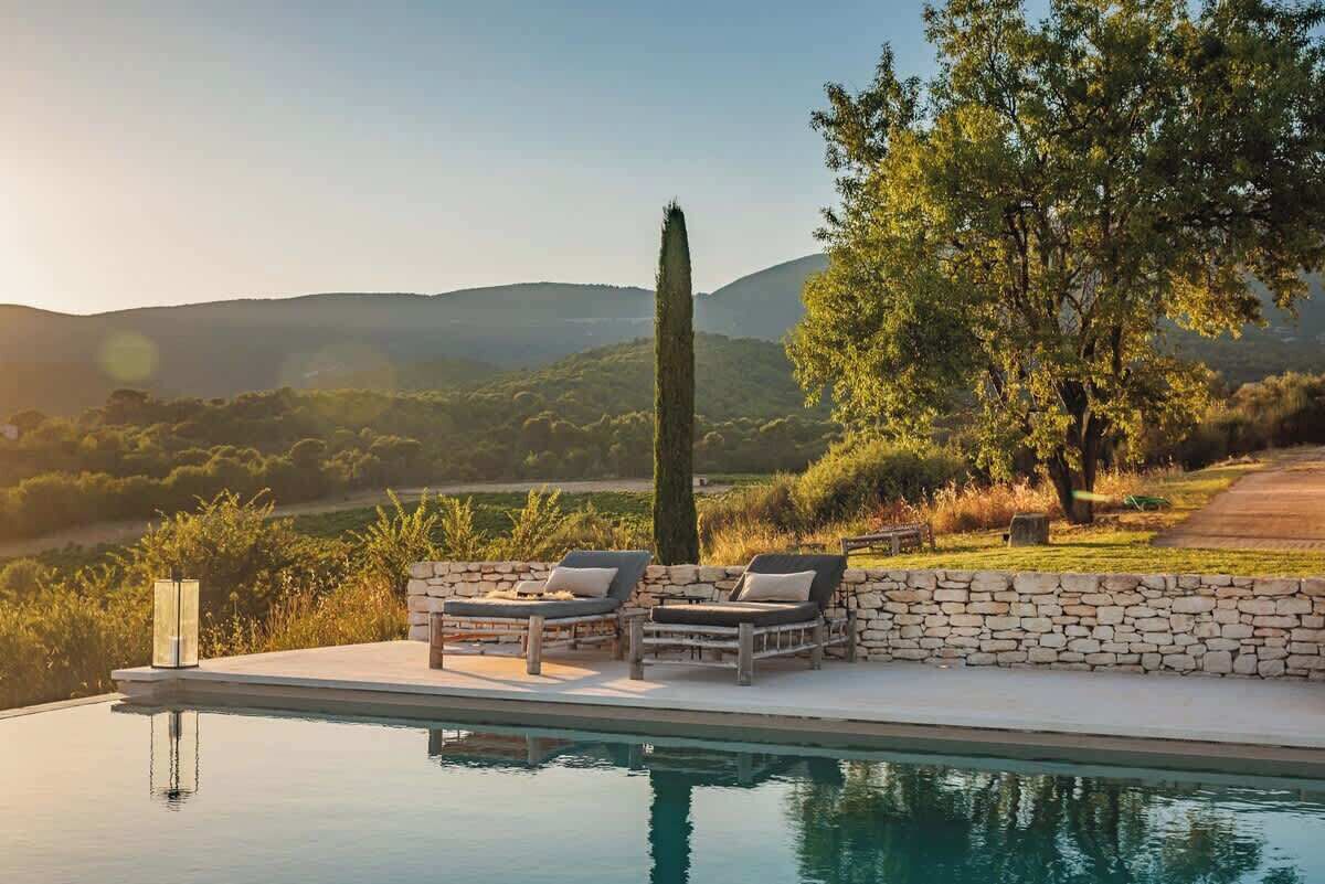 A poolside area with lounge chairs and a stone wall, surrounded by greenery and hills.