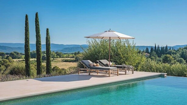 A poolside area with lounge chairs and an umbrella, surrounded by tall trees and a view of distant hills.