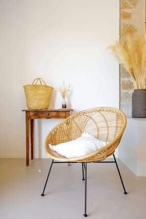 A wicker chair with a white cushion, a wicker basket on a wooden table, and a vase with dried plants on the wall.