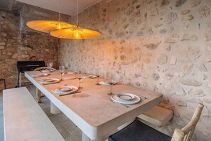 Uma longa mesa de jantar posta com pratos, copos e talheres, com uma parede de pedra e um lustre ao fundo.