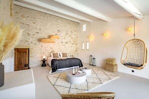 A bedroom with a stone wall, a bed with a black and white bedspread, a hanging chair, and a round coffee table. - Lourmarin, Htd pool A/C By the"DomainedelaColline" (Cabrières-d’Aigues)