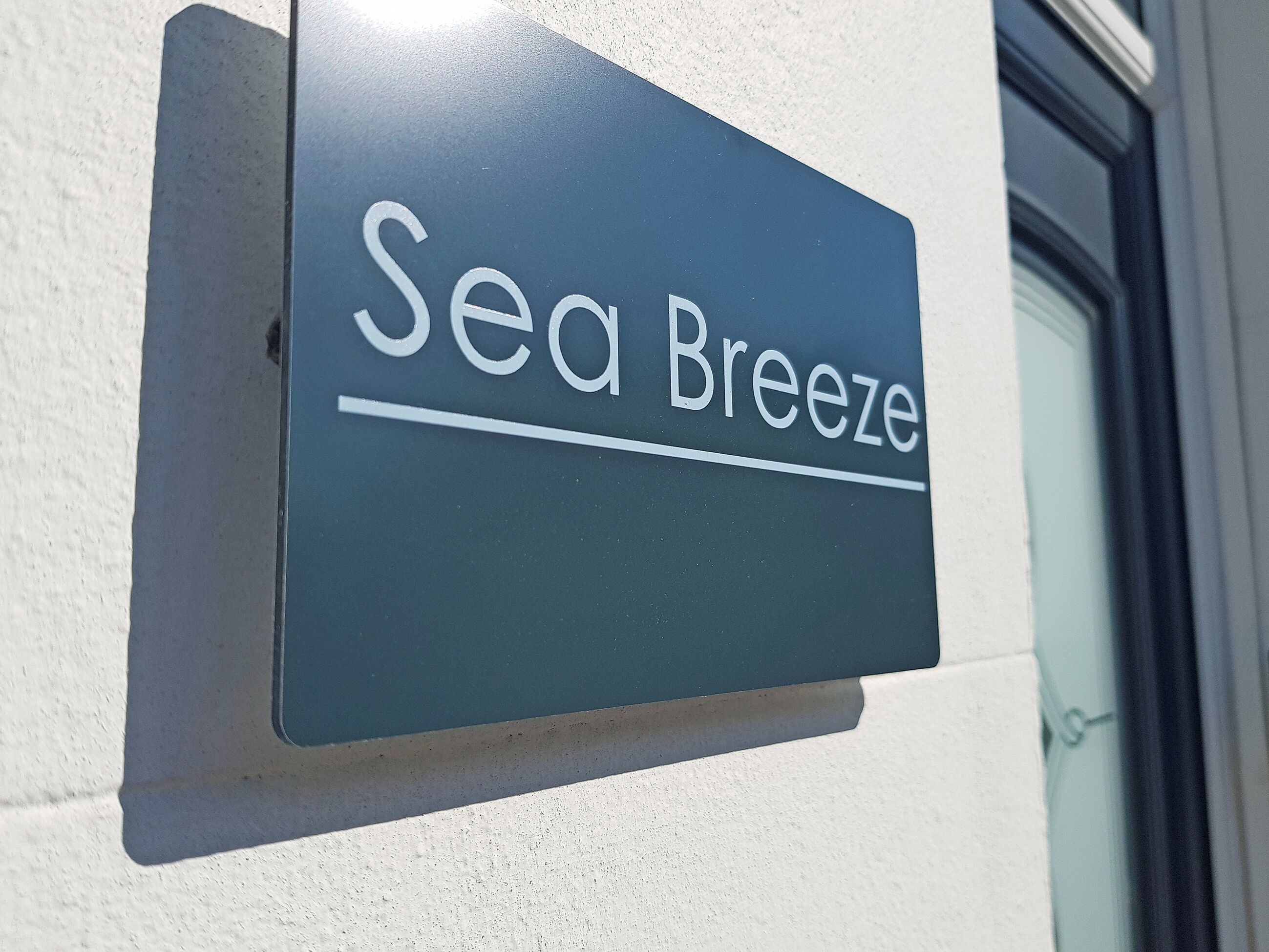 Et skilt med teksten "Sea Breeze" på mørk baggrund, monteret på en lys væg.