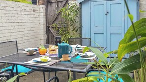 Una mesa de patio con mantel azul, rodeada de sillas y varios objetos como una tetera, tazas y comida.