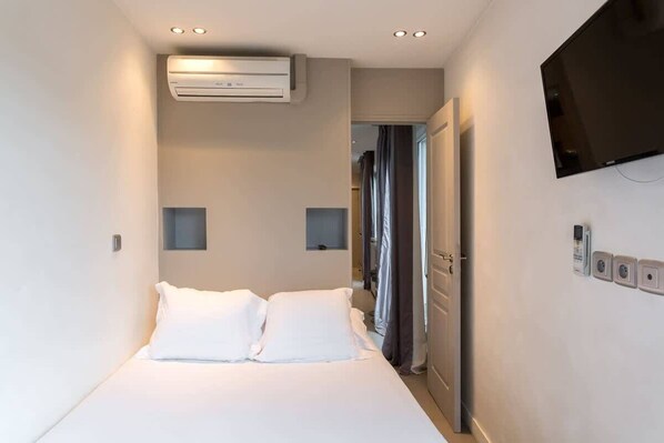 A hotel room with a bed, an air conditioning unit, a television mounted on the wall, and a door leading to a bathroom.