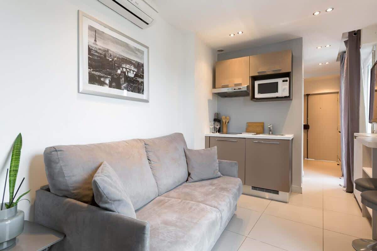 Un salon moderne avec un canapé gris, une petite table et une plante en pot. On y trouve également une kitchenette équipée d’un four à micro-ondes et de placards. Un tableau encadré est accroché au mur.