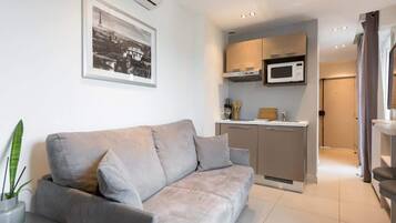 Un salon moderne avec un canapé gris, une petite table et une plante en pot. On y trouve également une kitchenette équipée d’un four à micro-ondes et de placards. Un tableau encadré est accroché au mur.