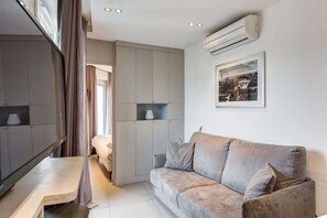 A modern, well-lit room with a grey sofa, a small table, and a bed in the background. There is a wall-mounted air conditioner and a framed picture above the sofa.