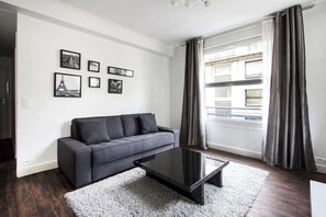 Una sala de estar moderna con un sofá gris, una mesa de centro de vidrio y un tapete blanco. Hay cuadros enmarcados en la pared y una ventana con cortinas.