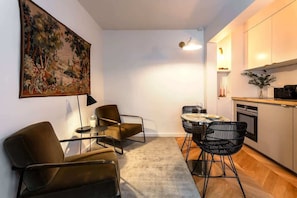 Una sala moderna con un sofá, una mesa de centro y una mesa de comedor con sillas. Hay un cuadro en la pared, una lámpara y una zona de cocina con armarios y encimera.