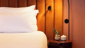 Cama bem arrumada com roupa de cama branca, abajur moderno e mesa de cabeceira pequena com objetos decorativos.