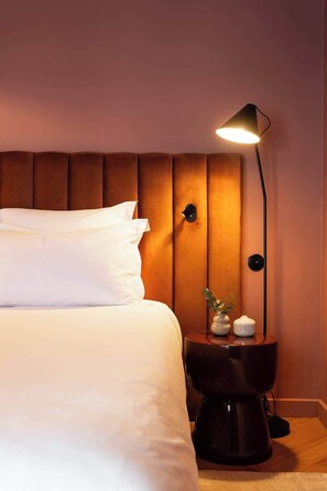 Un lit bien fait, avec du linge de lit blanc, une lampe de chevet moderne et une petite table de chevet agrémentée d’objets décoratifs.