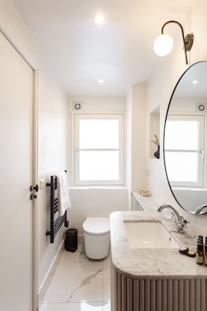 Une salle de bain moderne avec des toilettes blanches, un lavabo doté d’un plan de travail en marbre, un grand miroir et une fenêtre laissant entrer la lumière naturelle.