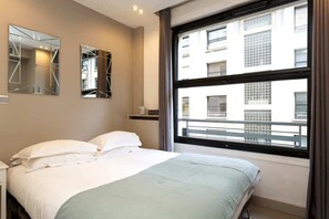 Habitación de hotel con cama, dos espejos en la pared, ventana con vista a edificios y un pequeño estante con un objeto decorativo.