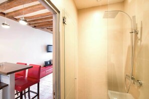A modern bathroom with a glass-enclosed shower, a wooden ceiling, and a red seating area visible through an open door.