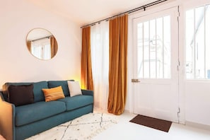 Ein Zimmer mit einem petrolfarbenen Sofa, einem runden Spiegel und einer weißen Tür mit Blick ins Freie.