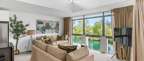 A modern living room with a sofa, a TV on a stand, a floor lamp, and a large window overlooking a body of water.