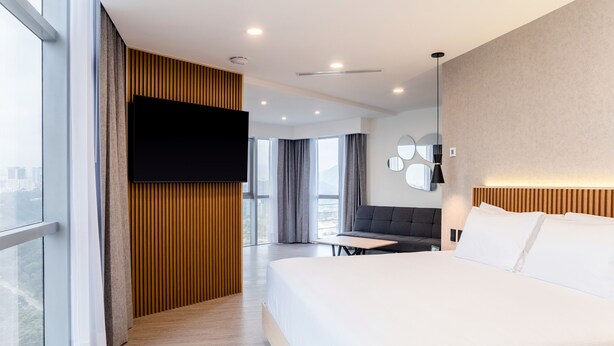 A modern hotel room with a large bed, a flat-screen TV mounted on a wooden panel, a seating area with a sofa and table, and a large window offering a city view. - HOTEL LUURE  (Huixquilucan)
